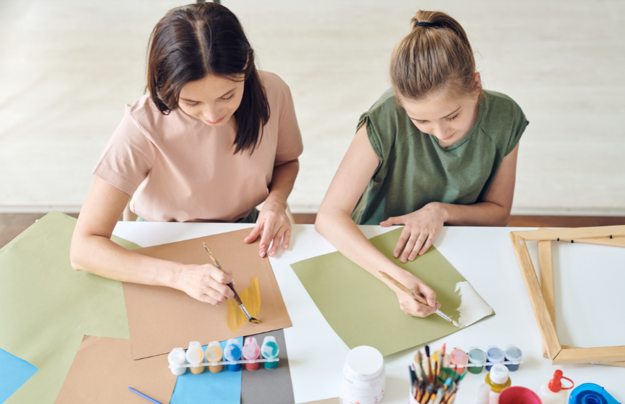 Young woman with her teenage daughter with paintbrushes painting watercolors on paper while sitting by the desk